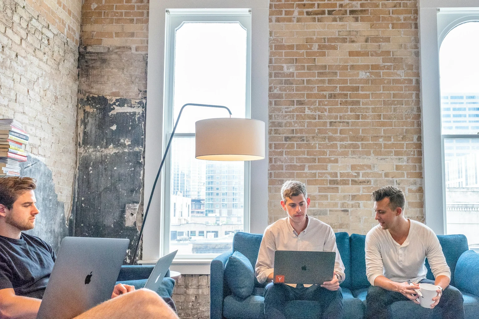 Three colleagues working on laptops on couches in a bright brick-walled office